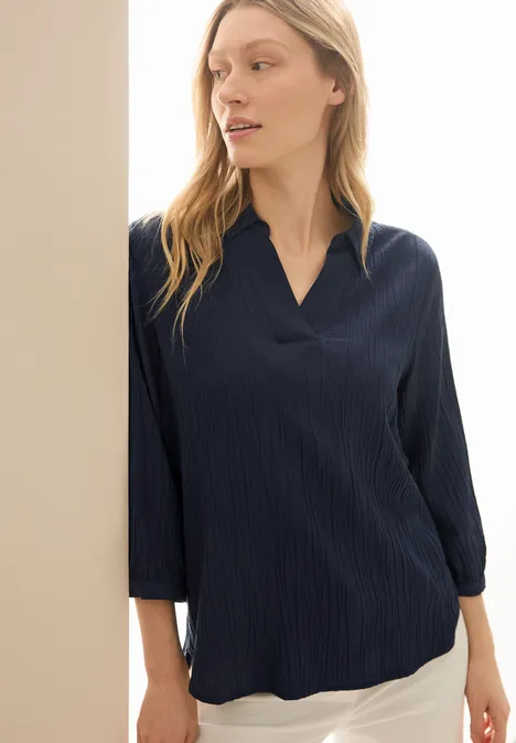 Frau mit dunkelblauer Bluse und weißen Hosen lehnt an einer Wand.
